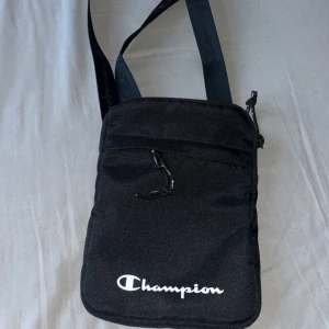 Svart axelremsväska från Champion - Säljer en stilren svart axelväska från Champion. Väskan har en justerbar axelrem och ett huvudfack med dragkedja, samt ett mindre fack på framsidan med dragkedja. Champion-loggan är tryckt i vitt på framsidan.