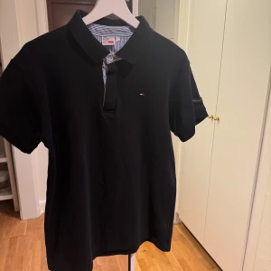 Svart pikétröja från Tommy Hilfiger - Snygg svart pikétröja från Tommy Hilfiger med klassisk krage och knappslå. Tröjan har en liten broderad logotyp på bröstet och randigt mönster på insidan av kragen. Perfekt för en stilren look till sommaren. Skick 8,5/10, storlek L sitter som M