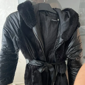Svart pälsjacka med bälte - Snygg svart jacka med pälsdetaljer och quiltad design. Jackan har en huva och ett bälte i midjan för en stilren look. Perfekt för kyligare dagar. Råkade beställa 2st av samma, därför är denna helt oanvänd!