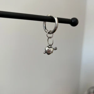 Silverörhänge i rostfritt stål med hjärthänge - Snyggt silverörhänge med ett litet hjärthänge. Perfekt för att ge en subtil men stilren touch till din look. Hängsmycket är format som ett hjärta med pil.