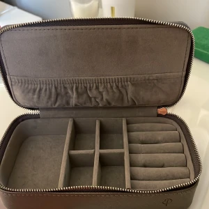 Grå smyckes skrin med dragkedja från Edblad - Snygg och praktisk grå smyckes skrin med dragkedja och flera fack för organisering. Perfekt för att hålla dina smycken säkra och organiserade. Skrinet har en stilren design med ett diskret märke på sidan.