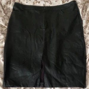 Svart skinnkjol med slits - Snygg svart skinnkjol med en stilren design och en diskret slits framtill. Kjolen har en knapp och dragkedja i midjan för enkel påklädning. Perfekt för att ge en edgy touch till din outfit. Obs! Den behöver strykas och jag uppfattar storleken som en 44. 