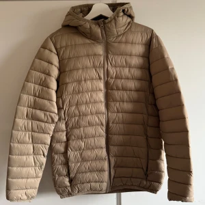 Beige pufferjacka från H&M - Säljer en snygg beige pufferjacka från H&M Divided. Jackan har en dragkedja framtill och en praktisk huva. Perfekt för kyligare dagar med sin quiltade design och långa ärmar. 