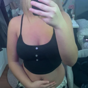 Svart croppad linnetopp - Säljer en svart croppad linnetopp med smala axelband och knappar framtill. Perfekt för en avslappnad stil.