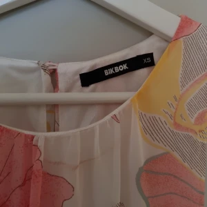 Blommig klänning från BikBok - Säljer en färgglad blommig klänning från BikBok i storlek XS. Klänningen har ett bälte i midjan och är ärmlös med ett vackert mönster i rosa, gult och grönt på en vit bakgrund. Perfekt för sommarens alla tillfällen!