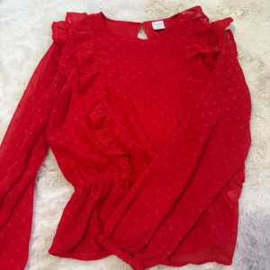 Röd blus från Lindex Kids  - Säljer en söt röd blus från Lindex med prickigt mönster och volangdetaljer. Blusen har långa ärmar och en elastisk midja för en bekväm passform. Den är i nyskick och har inte används. Perfekt för att liva upp garderoben för lite festligare tillfällen kanske. Den är i storlek 140. 