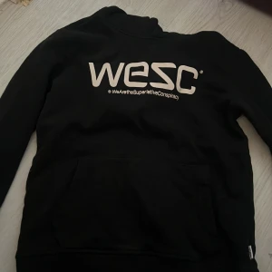 Svart hoodie från WESC - Säljer en svart hoodie från WESC med tryck på framsidan. Den har en klassisk design med huva och en stor ficka framtill. Perfekt för en avslappnad stil.