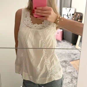 Vit ärmlös blus med spetsdetaljer - Säljer en söt vit blus med spetsdetaljer runt halsringningen. Blusen är ärmlös och har en luftig passform. Toppen har lite slitningar men säljer för ett bra pris!