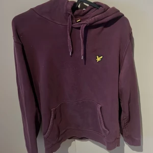 Lila hoodie från Lyle & Scott - Säljer en snygg lila hoodie från Lyle & Scott med en gul logga på bröstet. Tröjan har en klassisk känguruficka och justerbar huva med dragsko. Perfekt för en avslappnad stil.