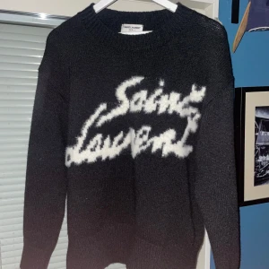 Svart stickad tröja från Saint Laurent - Säljer en svart stickad tröja från Saint Laurent med vit logotyp på framsidan. Tröjan har långa ärmar och en rund halsringning. Perfekt för en stilren och trendig look.