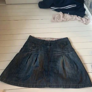 Jeanskjol från Spirella Denim - Snygg jeanskjol från Spirella Denim i en klassisk blå färg. Kjolen har en normal passform med dekorativa sömmar och knappar framtill. Insidan är fodrad med ett blommigt mönster som ger en extra touch. Perfekt för en avslappnad stil.