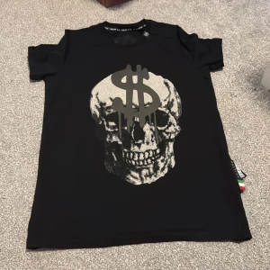 Svart t-shirt med dödskallemotiv qp  - Snygg svart t-shirt från Philipp Plein med ett stort dödskallemotiv och dollartecken på framsidan. T-shirten har en normal passform och är kortärmad, perfekt för en cool och avslappnad stil.