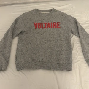 Grå sweatshirt från Zadig & Voltaire - Snygg grå sweatshirt från Zadig & Voltaire med röd text på framsidan. Perfekt för en avslappnad stil. Tröjan har långa ärmar och en rund halsringning.