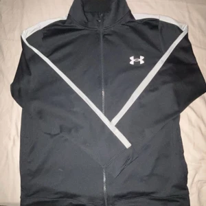 Svart tröja från Under Armour - Säljer denna snygga svart träningströja/ jacka från Under Armour med vita detaljer. Tröjan  har en dragkedja framtill och loggan på bröstet. Perfekt för träning eller en sportig look. Den är knappt använd.