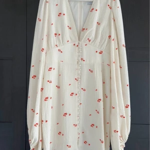Vit blommig klänning från ADOORE - Säljer en söt vit klänning från ADOORE med små röda blommor. Klänningen har en v-ringad design och knappar framtill. Den har långa ärmar med en lätt puffig stil och är perfekt för sommarens tillfällen. 🌸i nyskick och passaren storlek 40 eller L
