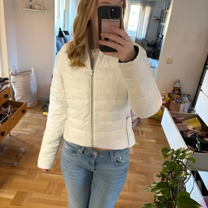 Vit pufferjacka med dragkedja - Säljer en stilren vit pufferjacka med dragkedja framtill. Jackan har långa ärmar och två praktiska fickor med dragkedjor. Perfekt för kyligare dagar och ger en snygg look.