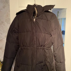 Svart dunjacka med huva - Säljer en svart dunjacka från JH Collection. Den är perfekt för vintern med sin varma och vadderade design. Jackan har en praktisk huva och stängs med dragkedja och knappar. Den har en normal passform och är långärmad, vilket gör den idealisk för kalla dagar.