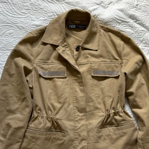 Beige jacka från Zara(safarijacka) - Säljer en snygg beige jacka (safarijacka) från Zara i storlek S. Jackan har långa ärmar och en klassisk krage. Den är dekorerad med glittriga detaljer på bröstfickorna och har en knappstängning framtill. Perfekt för vår och höst!