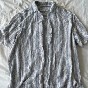 Grå och vit randig skjorta från Zara - Säljer en snygg och bekväm grå och vit randig overshirt från Zara. Overshirten är kortärmad och har knappar framtill. Perfekt för sommarens varma dagar eller en avslappnad vårdag. Materialet känns lätt och luftigt, vilket gör den idealisk för både vardag och strandhäng. Frågor? Skriv!