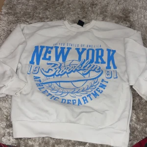 Vit sweatshirt från Gina Tricot - Säljer en vit sweatshirt från Gina Tricot med tryck i blått. Texten 'New York Brooklyn Athletic Department' ger en sportig känsla. Tröjan är i storlek XS och har långa ärmar. Perfekt för en avslappnad stil!