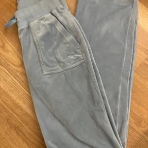Grå mjukisbyxor med fickdetaljer - Säljer ett par superbekväma ljusblå juciy couture mjukisbyxor. De har en elastisk midja och praktiska fickor med knappdetaljer. Perfekta för en avslappnad dag hemma eller en snabb tur till affären. Materialet känns mjukt och skönt mot huden.