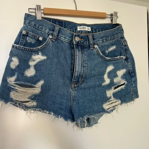 Blå jeansshorts från Pull&Bear - Säljer ett par snygga blå jeansshorts från Pull&Bear i storlek S. De har en cool, sliten look med fransiga kanter och flera slitningar fram. Perfekta för sommaren och ger en avslappnad stil. Hårt material.