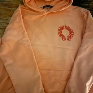 Ljus rosa hoodie från Chrome Hearts - Säljer en snygg orange hoodie från Chrome Hearts med deras ikoniska logga i rött på bröstet. Perfekt för en avslappnad stil med en touch av lyx. Hoodien har en klassisk passform och är tillverkad i mjukt material för extra komfort.