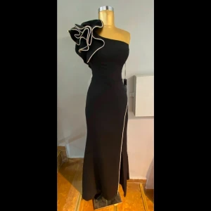 Balklänning: Svart one-shoulder klänning med dramatisk silvrig volangdetalj - Klänningen är i srl 40 dock skräddarsydd att passa någon i storlek 36/S! Säljer denna eleganta svarta långklänning med en one-shoulder design och en iögonfallande volangdetalj på axeln. Klänningen har endast används en gång och är köpt i Turkiet💕pris kan diskuteras