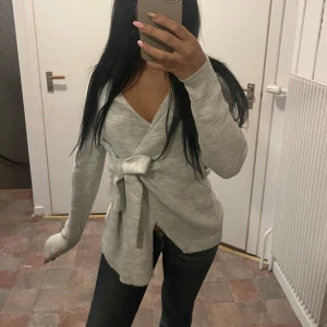 Grå omlottkofta - Säljer en stilren grå omlottkofta med knytband i midjan. Koftan har långa ärmar och en v-ringad design som ger en elegant look. Perfekt för kyligare dagar.