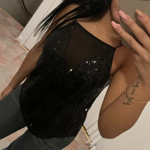 Svart glittrig ärmlös topp - Snygg svart glittrig topp med transparent mesh-detalj vid halsen. Toppen är ärmlös och har en elegant design som passar perfekt för festliga tillfällen. Den har en smickrande passform och tunna axelband.