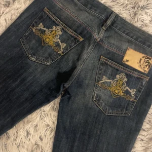 Mörkblåa Diesel jeans - Snygga mörkblå, lågmidjade, bootcut jeans från Diesel med unika broderade detaljer på bakfickorna. Jeansen har en klassisk femficksdesign och kontrastsömmar i rött. Perfekta för en stilren look. Står storlek 29/32 men jag själv är st 27-28 i midjan och de är för små så skulle säga att de sitter som 26/32 istället. Priset kan diskuteras vid snabb affär!