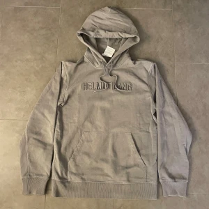 Grå hoodie från Helmut Lang - Säljer en stilren grå hoodie från Helmut Lang med broderad logga på bröstet. Tröjan har en klassisk känguruficka och justerbar huva med snören. Perfekt för en avslappnad look. Pris ej hugget i sten!
