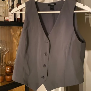 Grå väst från Lindex - Snygg grå väst från Lindex med knappar framtill. Den har en klassisk v-ringad design och passar perfekt för en stilren look. Västen är i en neutral grå färg som är lätt att matcha med andra plagg. Aldrig använd.