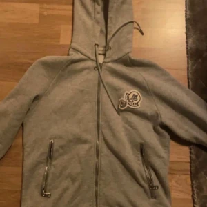 Grå hoodie från Moncler - Säljer en snygg grå hoodie från Moncler med dragkedja och huva. Den har ett broderat Moncler-märke på bröstet och praktiska fickor med dragkedja. Perfekt för en casual look. 9/10 skick. 