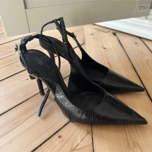 Svarta pumps med spetsig tå - Eleganta svarta pumps med spetsig tå och hög klack. Skorna har en textur som liknar ormskinn och justerbara remmar med guldfärgade spännen. Perfekta för en stilfull look.