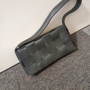 Svart axelväska från NLY Accessories - Snygg svart axelväska i flätad design från NLY Accessories. Väskan har en praktisk dragkedja inuti och stängs med tryckknappar. Perfekt för att bära dina nödvändigheter med stil.