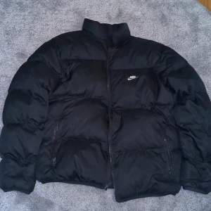 Nike svart pufferjacket  - Säljer en svart pufferjacka från Nike med dragkedja och broderad logga på bröstet. Jackan har långa ärmar och är perfekt för kyligare dagar. Den är både snygg och funktionell. Säljer för att jag behövder cash