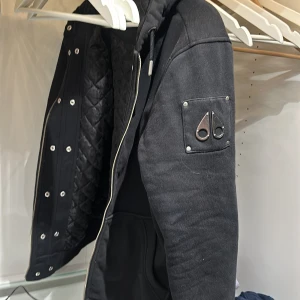 Svart jacka från Moose Knuckles - Snygg svart jacka från Moose Knuckles med quiltad insida och dragkedja. Jackan har en cool logga på ärmen och knappar framtill. Perfekt för kyligare dagar. Nypris 3700 , original bag samt pälsen som tillhör finns.
