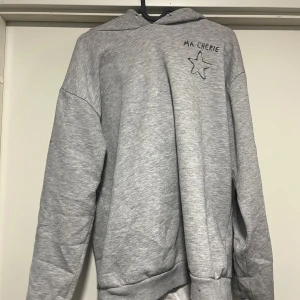 Grå hoodie - Säljer en grå hoodie med texten 'MA CHERIE' och en stjärna på bröstet. Ärmen har flera stjärnor tryckta längs sidan. Perfekt för en avslappnad stil.