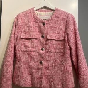 Rosa vårjacka - Säljer en stilren rosa jacka med rund hals och dekorativa knappar framtill. Kavajen har en klassisk passform med långa ärmar och två fickor med lock. Perfekt för en chic look!