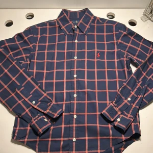 Rutig skjorta från Ralph Lauren - Snygg rutig skjorta från Ralph Lauren i marinblått med röda och vita rutor. Skjortan har långa ärmar och knappar framtill. Den är broderad med det klassiska Ralph Lauren-logotypen på bröstet. Perfekt för en stilren look! Måtten på skjortan är längd 65 cm, midje bredd 50 cm, axelbredd 45 cm.