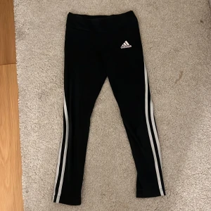Svarta leggings från Adidas - Säljer ett par svarta leggings från Adidas med de klassiska tre vita ränderna längs sidorna. Perfekta för träning eller en avslappnad dag. De är tillverkade i ett stretchigt syntetmaterial för bästa komfort.