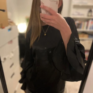 Svart blus med volangärmar - Elegant svart blus med långa volangärmar i ett glansigt silkesmaterial. Perfekt för ett speciellt sammanhang t.e.x. Fest eller nyår🩷. Blusen har en rund halsringning och en lös passform. Blusen har storleken 158-164 och har används ett fåtal gånger. Det finns inga tecken på att den har används utan är nästan i nyskick🫶🏼