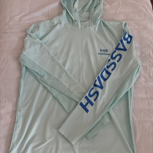 Ljusblå fishing hoodie från Bassdash - Säljer en ljusblå hoodie från Bassdash med lång ärm och tryck på ärmen. Tillverkad i polyester och spandex för en bekväm passform. Perfekt för utomhusaktiviteter med UPF 50+ UV-skydd.