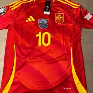 Röd Spanien fotbollströja med nummer 10 - Snygg röd Spanien fotbollströja från Adidas med gula detaljer och nummer 10 på framsidan. Tröjan har korta ärmar och är tillverkad i Aeroready-material för optimal komfort. Perfekt för fotbollsfans som vill visa sitt stöd för laget.