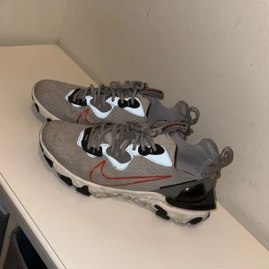 Grå Nike sneakers med orange detaljer - Snygga grå Nike sneakers med orange swoosh och svarta detaljer. Skorna har en modern design med snörning och en bekväm sula. Perfekta för en sportig look.