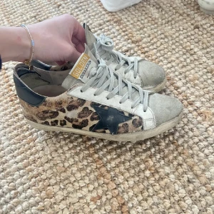 Leopardmönstrade sneakers från Golden Goose - Snygga sneakers från Golden Goose med leopardmönster och en svart stjärndetalj på sidan. Skorna har snörning och en beige bas med svarta detaljer vid hälen. Perfekta för en trendig look. Skorna är någorlunda slitna och sulan e lite nersplibad i hällen, kan skicka bilder vid önskan