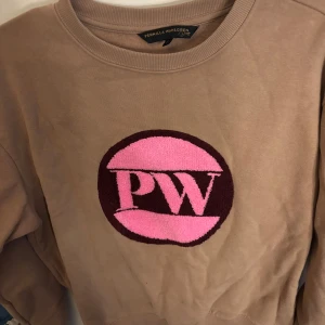 Beige tröja från Pernilla Wahlgren - Säljer en beige tröja från Pernilla Wahlgren med ett stort rosa och vinrött PW-märke på framsidan. 