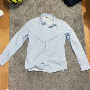 Ljusblå randig skjorta från H&M - Snygg ljusblå skjorta från H&M med smala ränder och små svarta detaljer. Skjortan har långa ärmar och knappar framtill. Perfekt för en stilren look.