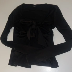 Svart långärmad topp från Gina Tricot - Säljer en stilren svart långärmad topp från Gina Tricot. Toppen har en knytning framtill som ger en snygg detalj och är även öppen vid hela magen. Perfekt för att matcha med olika outfits. Hör av dig vid intresse!